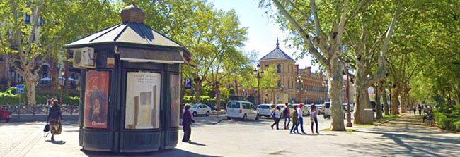 Un kiosco con publicidad y gente en una avenida de Sevilla